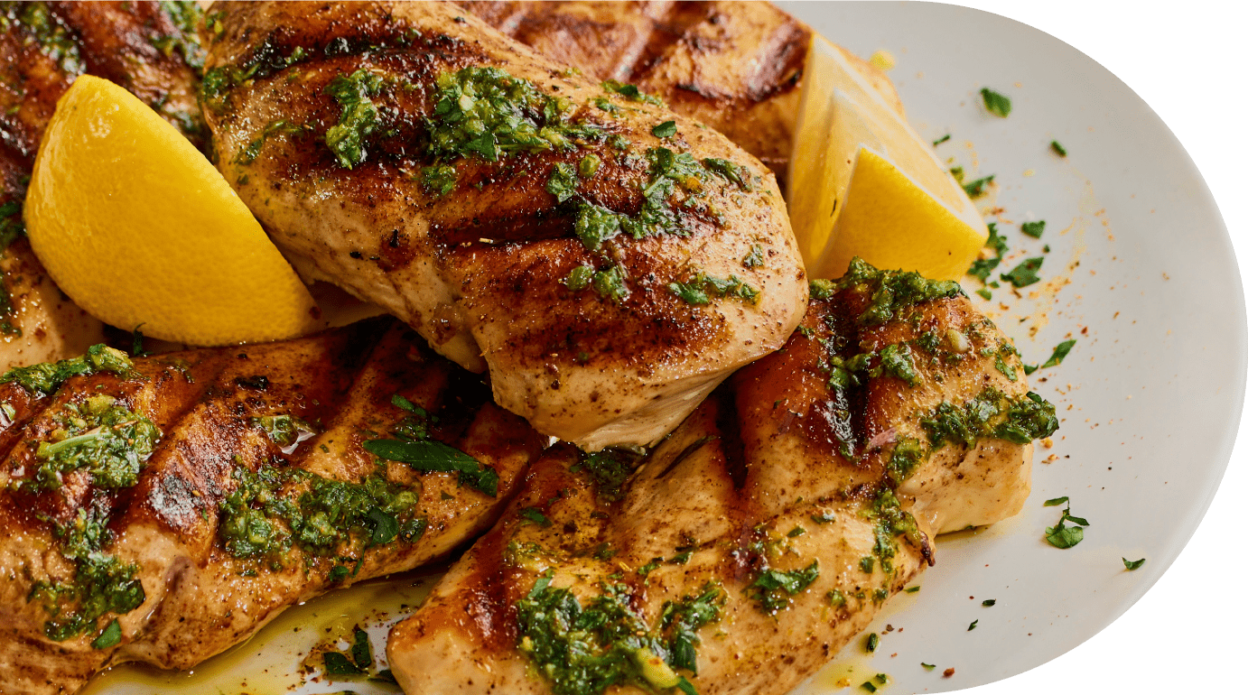 Cater Chicken - Grilled chicken breasts topped with green herb sauce, garnished with chopped herbs and served with lemon wedges on a white plate.
