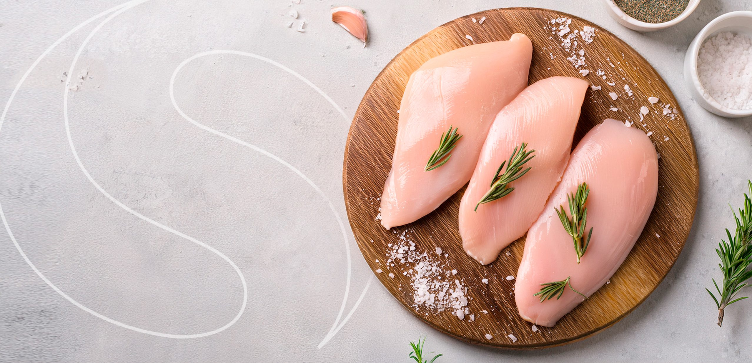 Three raw chicken breasts with sprigs of rosemary on a round wooden board, surrounded by coarse salt, pepper, and rosemary on a light gray surface.