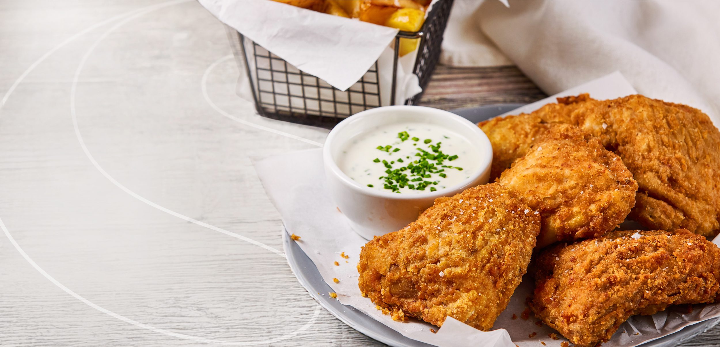 A plate of crispy fried chicken pieces with a small bowl of creamy dipping sauce topped with chopped herbs, next to a basket of French fries on a white surface.
