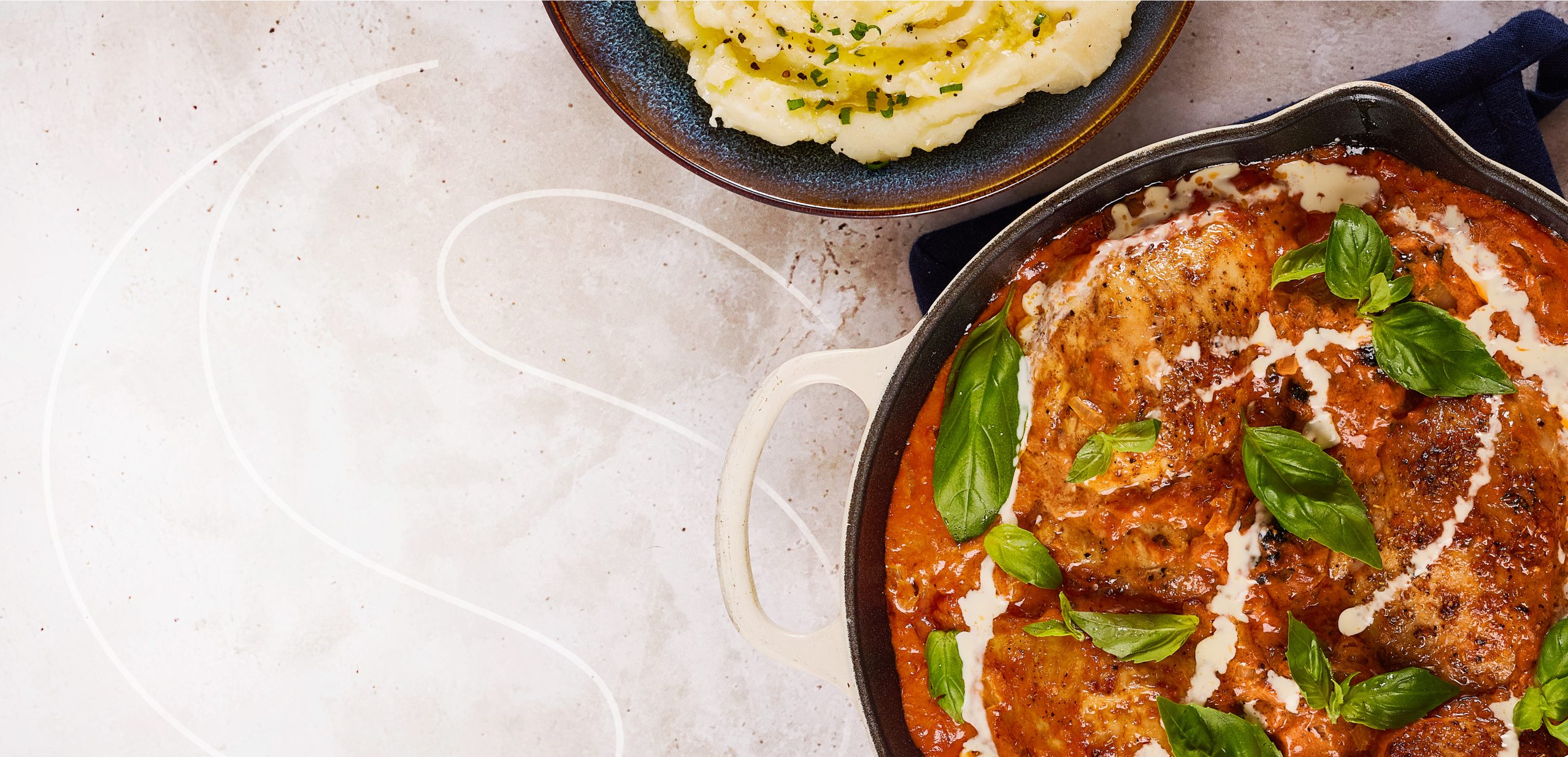A skillet of creamy tomato basil chicken garnished with fresh basil leaves, next to a bowl of mashed potatoes topped with herbs, on a light-colored surface.