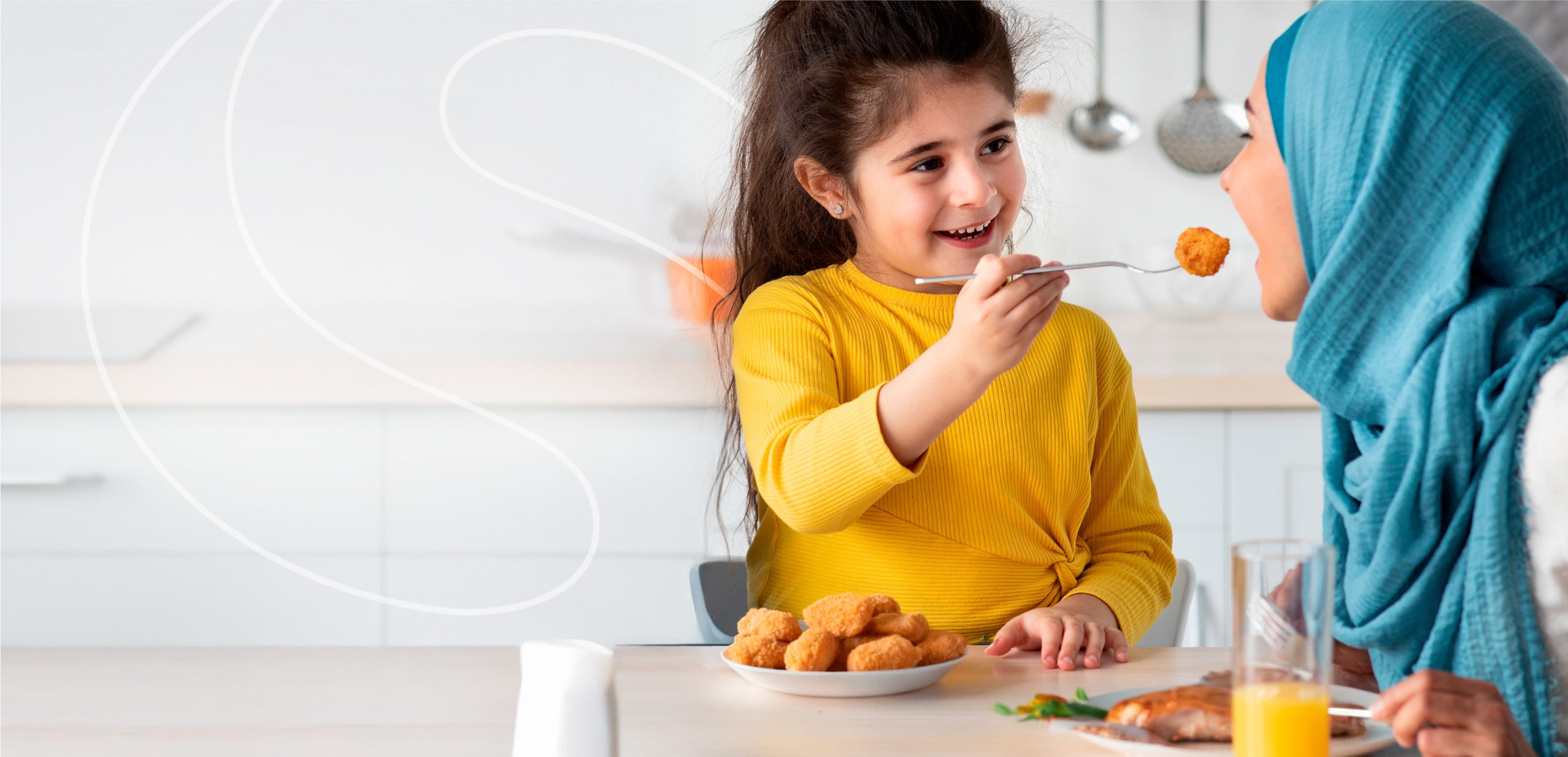 A smiling young girl in a yellow shirt feeds a woman in a blue headscarf a nugget at a kitchen table, with a plate of nuggets and a glass of orange juice in front of them.