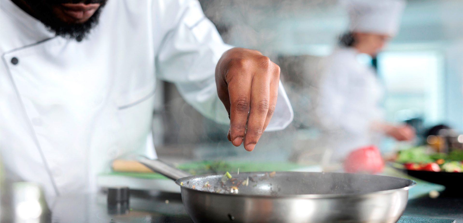 A chef in a white uniform sprinkles seasoning into a steaming pan, while another chef works in the blurred background of a professional kitchen.